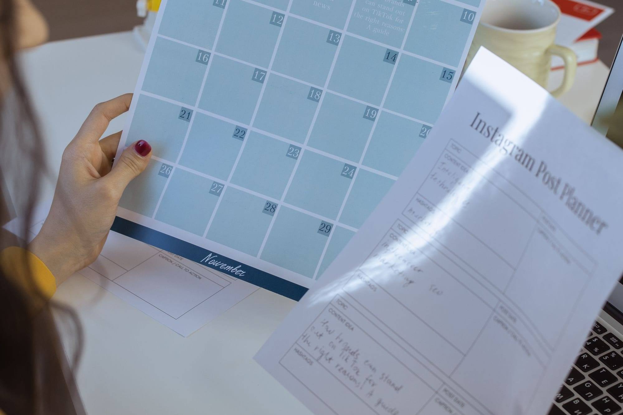 Manos que sostienen un calendario mensual de contenido y una plantilla para planificar publicaciones de Instagram junto a un portátil
