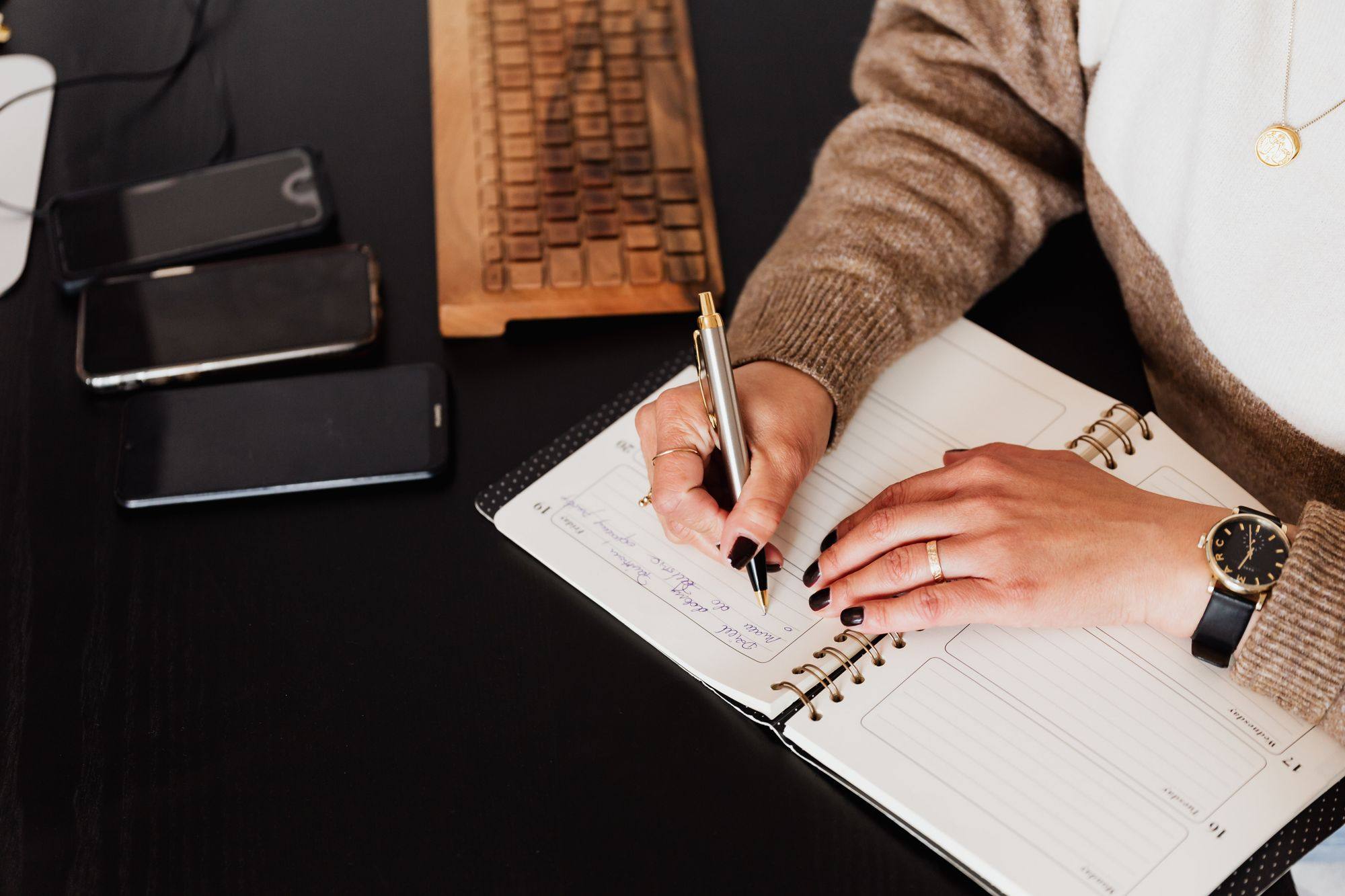 Manos de una mujer escribiendo en una agenda abierta sobre un escritorio negro, junto a tres teléfonos y un teclado de madera