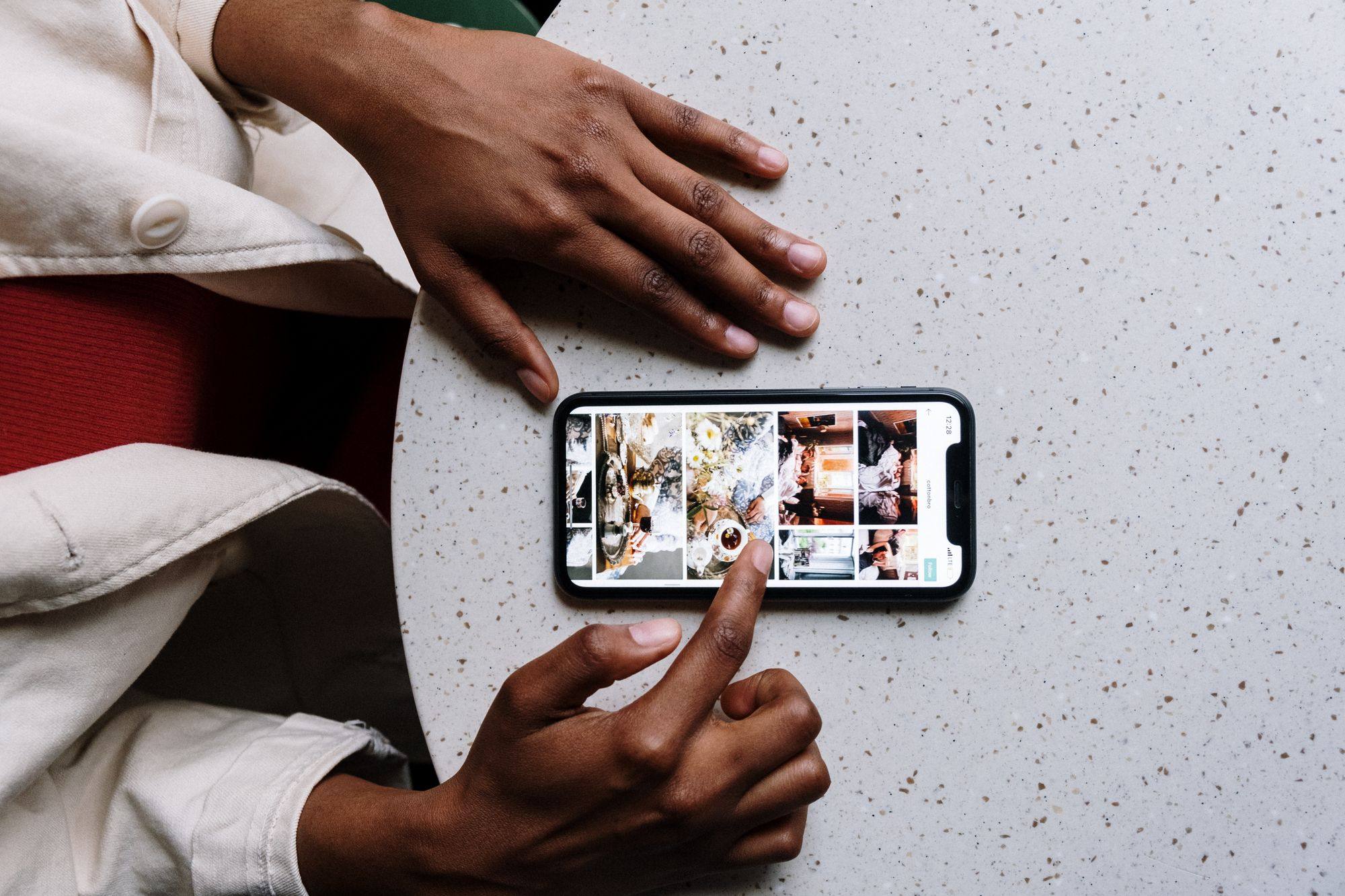 Manos sobre una mesa clara junto a un móvil que muestra una cuadrícula de fotos de un perfil, con un dedo a punto de tocar la pantalla