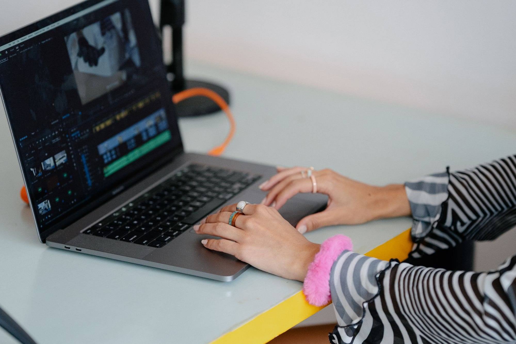 Manos editando un vídeo en un portátil sobre una mesa blanca y amarilla, con un micrófono de estudio al fondo